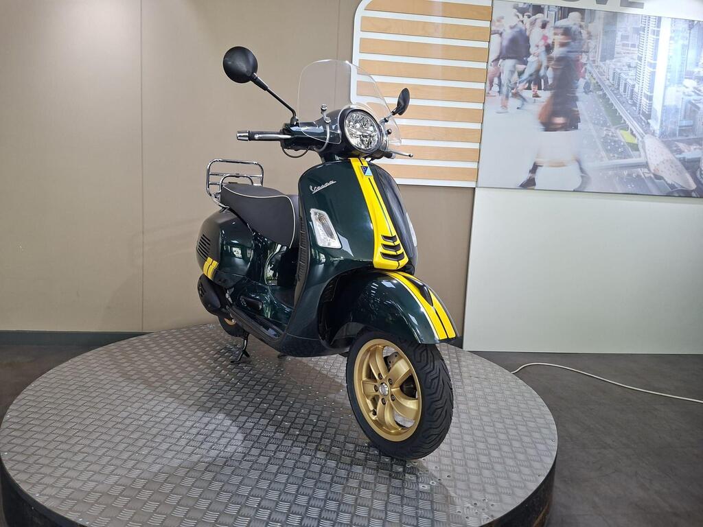 Vespa GTS 300 Racing Sixties 2022 4 A vintage-style Vespa scooter in dark green with yellow accents, displayed on a round metallic platform. The backdrop features a blurred image of a busy street scene.