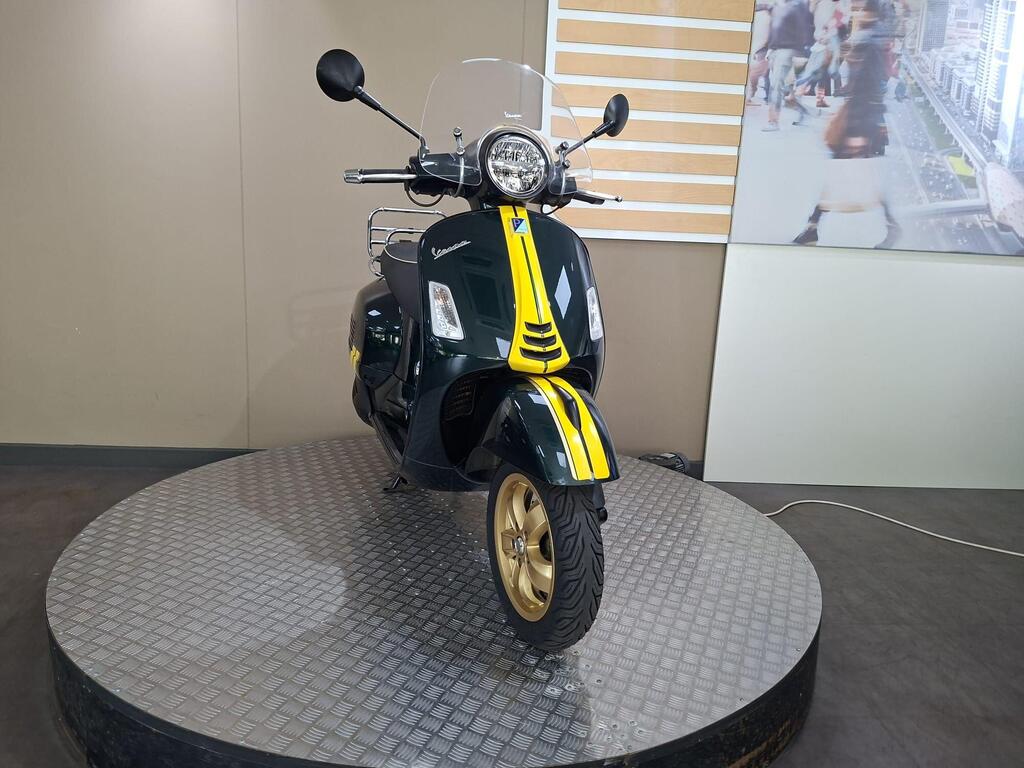 Vespa GTS 300 Racing Sixties 2022 5 A green Vespa scooter with yellow accents is displayed on a circular metal platform in a showroom setting, featuring a clear windshield and a decorative backdrop.