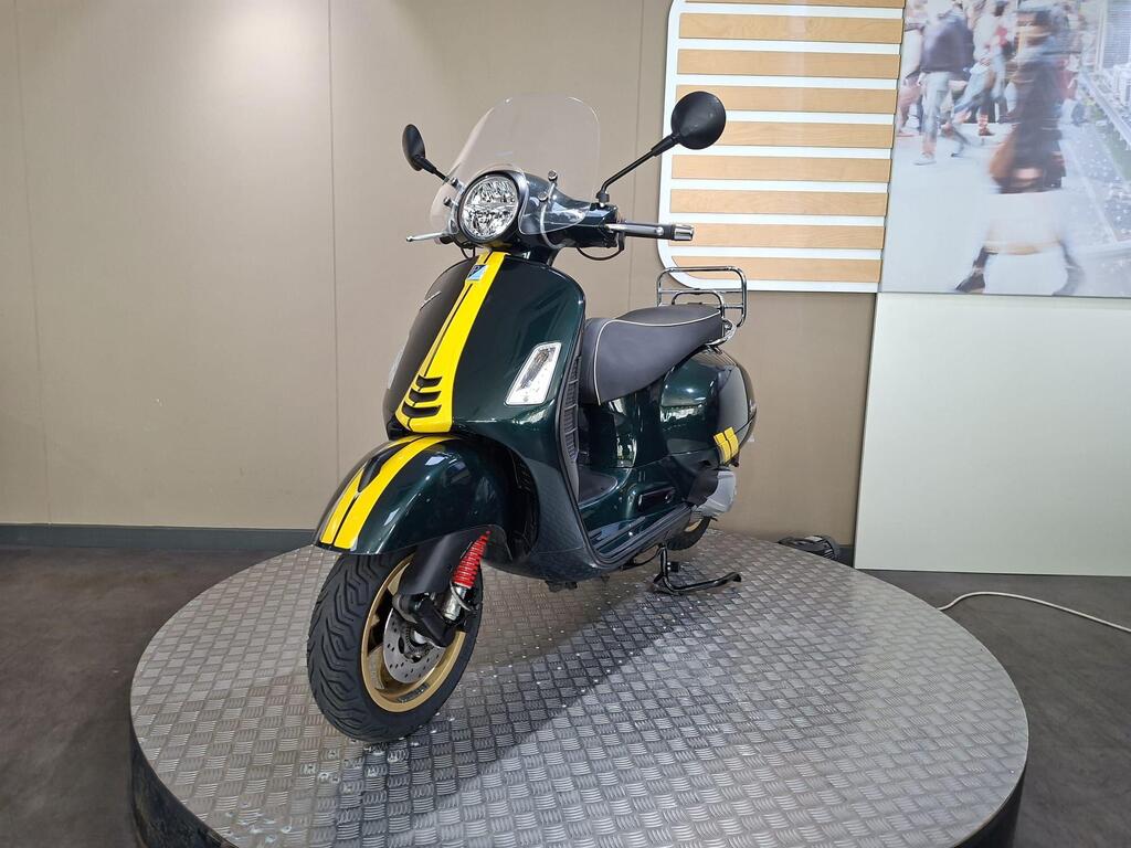 Vespa GTS 300 Racing Sixties 2022 6 A green scooter with yellow stripes and a windscreen, displayed on a circular metallic platform in a modern exhibit setting.