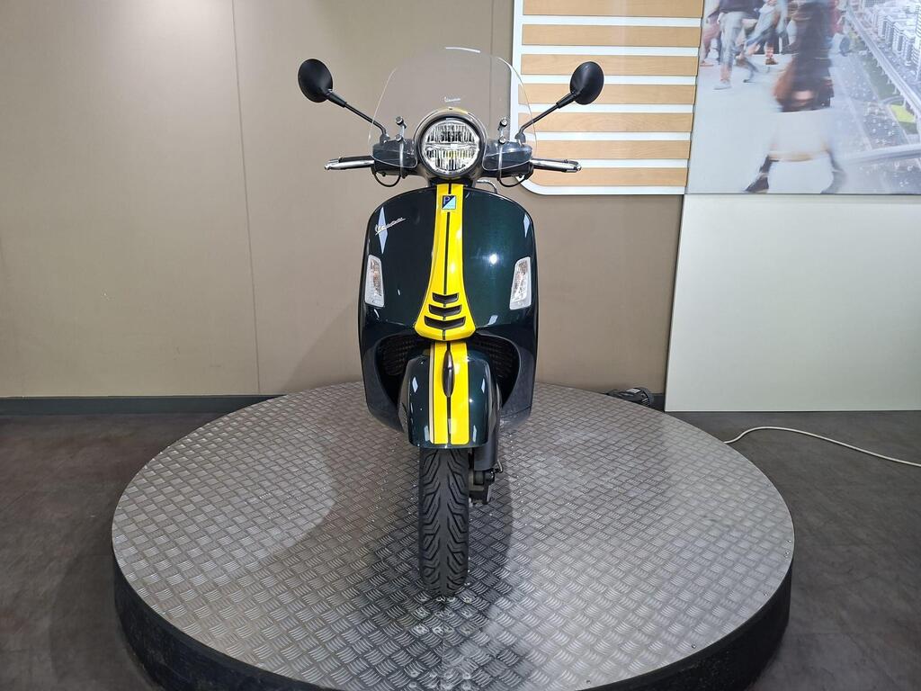 Vespa GTS 300 Racing Sixties 2022 7 A front view of a green and yellow scooter on a circular display stand, with a transparent windscreen and chrome accents. The background features a neutral wall and a blurred image of a bustling street.