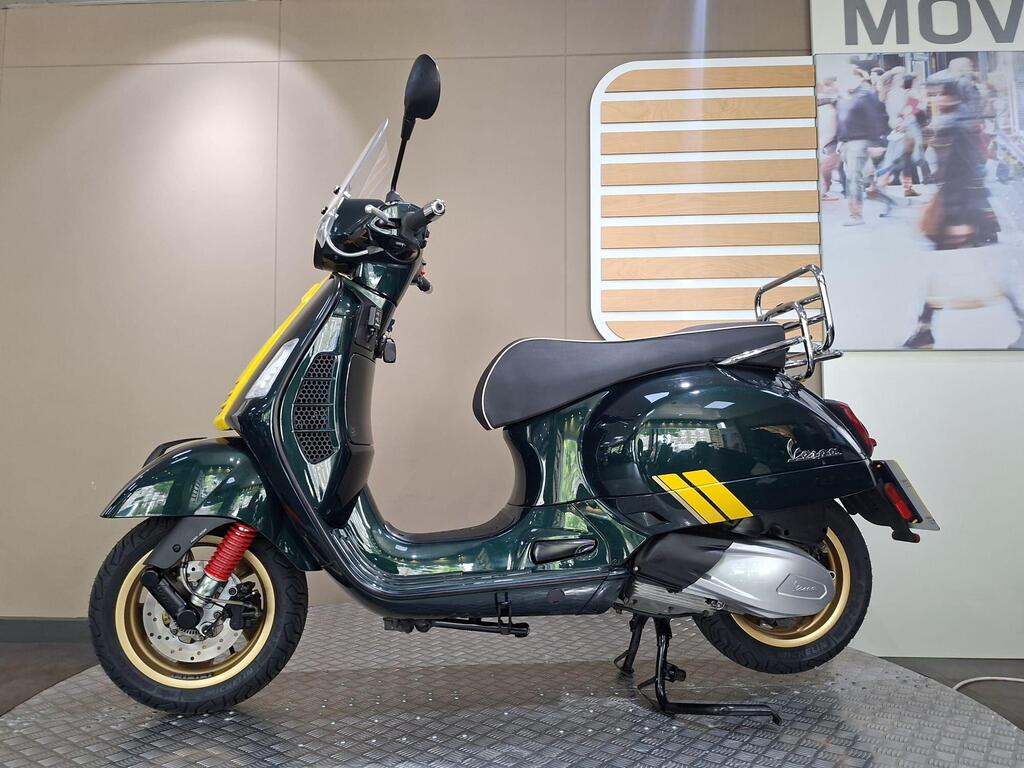 Vespa GTS 300 Racing Sixties 2022 8 A green Vespa scooter with yellow accents, featuring a shield, rear rack, and red suspension, displayed on a patterned floor against a neutral background.