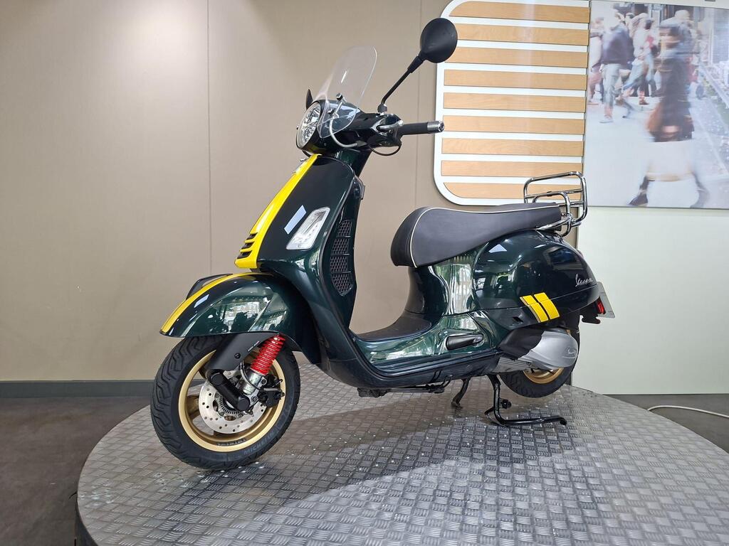 Vespa GTS 300 Racing Sixties 2022 9 A green Vespa scooter with yellow accents and a clear windscreen is displayed on a circular stand against a neutral-colored wall.