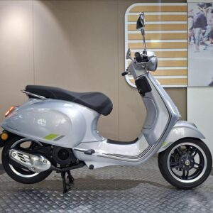 Vespa Primavera 125 2025 A silver scooter is displayed in profile against a neutral backdrop with a graphic of a busy street scene in the background. The scooter features a sleek design with a black seat, chrome accents, and distinctive wheels.