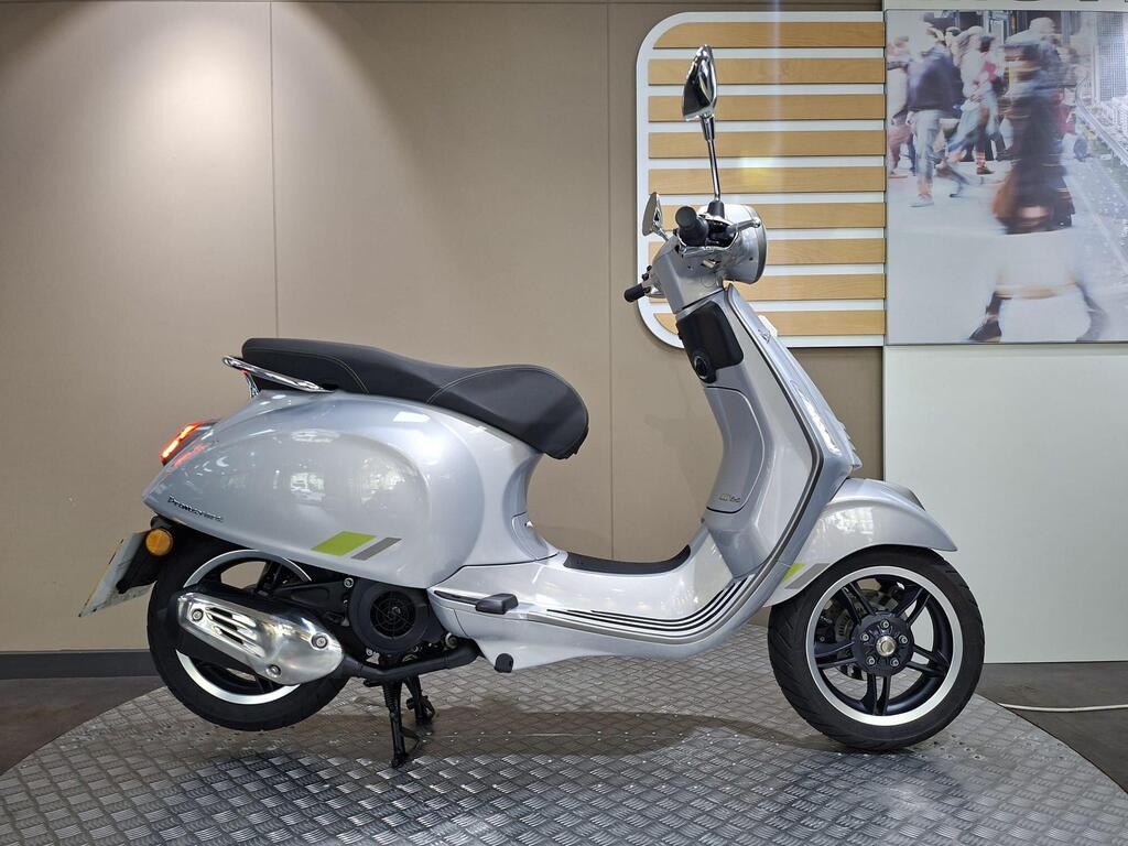 Vespa Primavera 125 2025 1 Vespa Primavera 125 2025 A silver scooter is displayed in profile against a neutral backdrop with a graphic of a busy street scene in the background. The scooter features a sleek design with a black seat, chrome accents, and distinctive wheels.