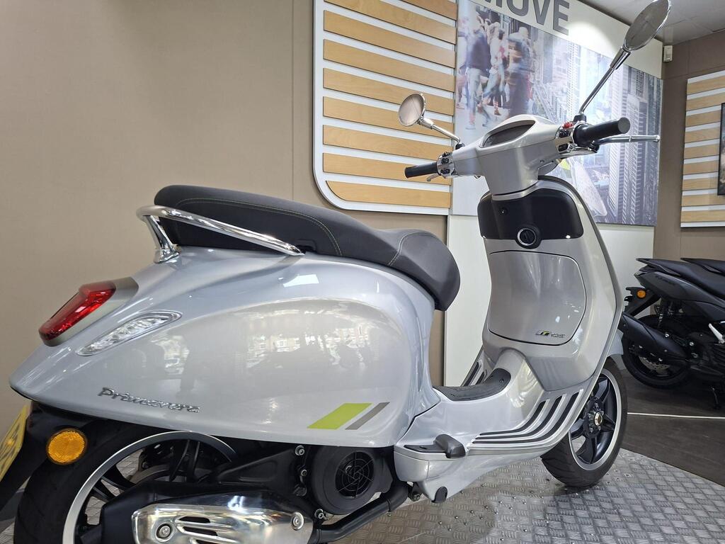 Vespa Primavera 125 2025 10 A silver scooter parked in a showroom with modern decor, featuring a sleek design and tinted mirrors. The floor is metallic, and a black scooter is visible in the background.