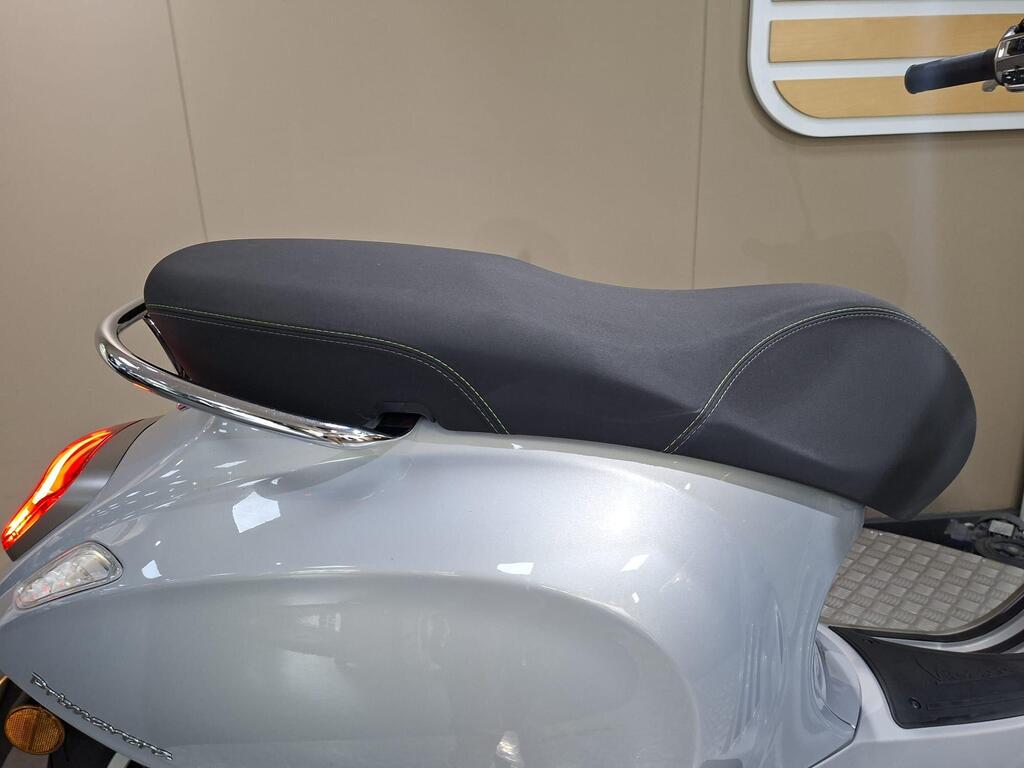 Vespa Primavera 125 2025 12 A close-up view of a sleek silver motorcycle seat with a black cover, featuring contrasting green stitching and a chrome rear grab rail. The background is a neutral beige.