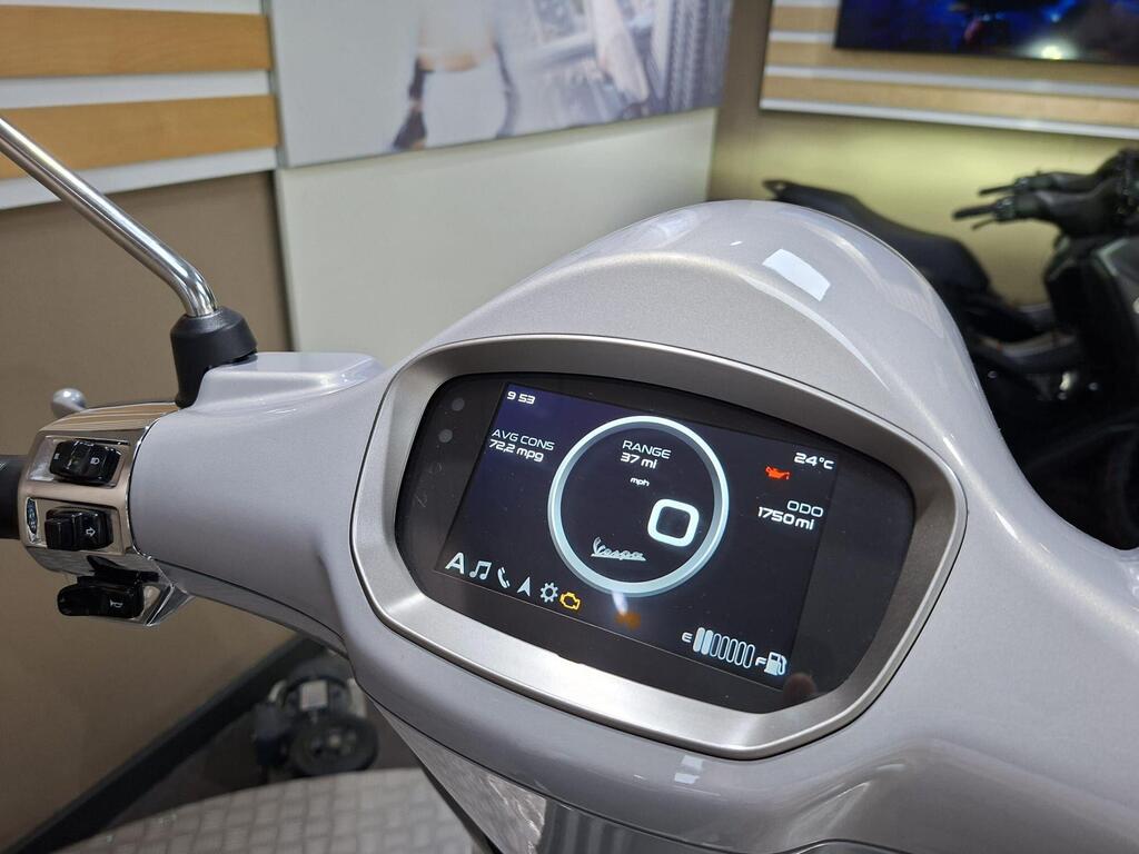 Vespa Primavera 125 2025 13 A close-up view of a scooter's dashboard displaying a digital screen with information including average consumption of 72.2 mpg, a range of 37 miles, current temperature of 24°C, and an odometer reading of 1750 miles. The dashboard is located on a light-colored scooter, with buttons and controls visible on the left side.