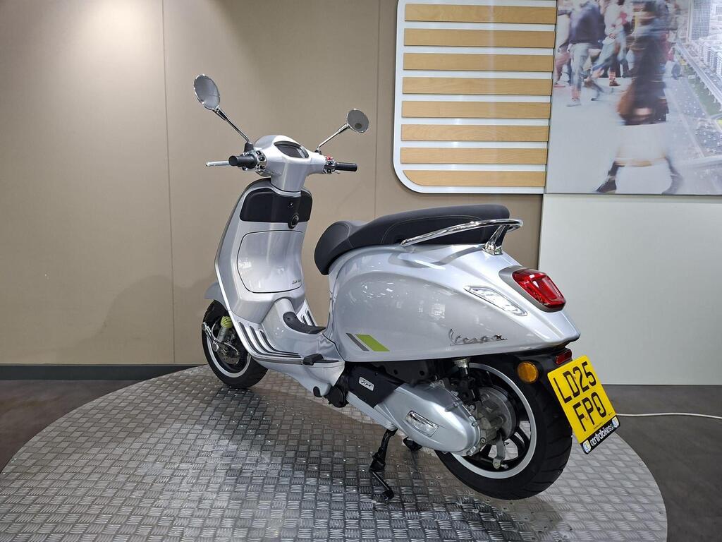 Vespa Primavera 125 2025 14 A silver Vespa scooter is displayed on a textured circular platform in an indoor showroom, with a modern backdrop featuring people and city imagery. The scooter is viewed from a three-quarter angle, highlighting its design and features.