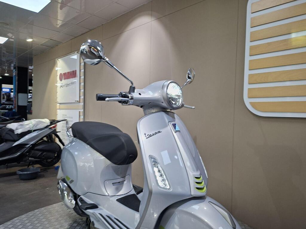 Vespa Primavera 125 2025 15 A silver scooter with a rounded front, featuring a prominent headlight and a rearview mirror, is displayed against a neutral-colored wall with a Yamaha logo and signage in the background.