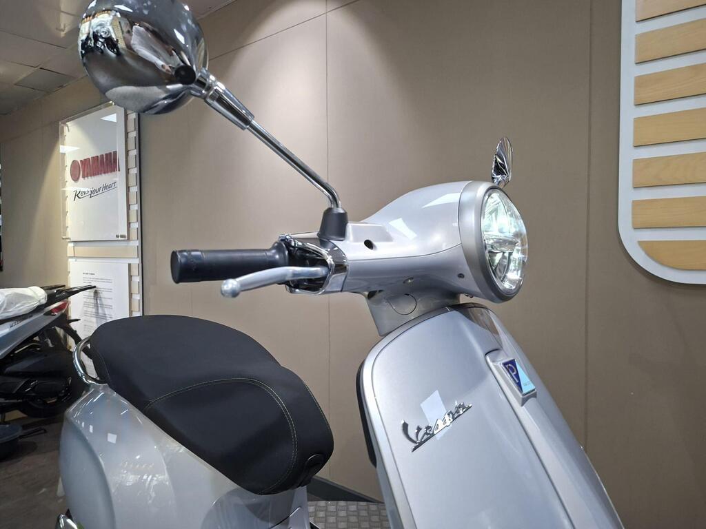 Vespa Primavera 125 2025 16 A close-up of a silver Vespa scooter with a round headlight, mirror, and handlebar against a neutral-colored wall.