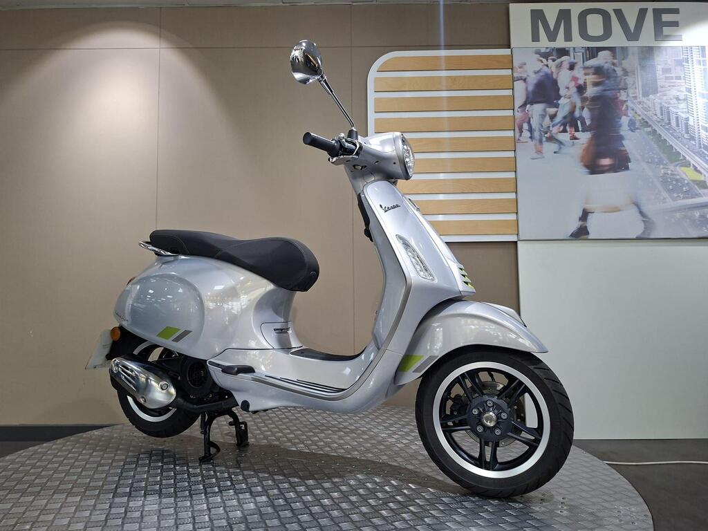 Vespa Primavera 125 2025 2 A silver Vespa scooter is showcased at an angle on a circular display platform, with a modern, minimalistic background featuring the word "MOVE" and an image of a bustling city scene.