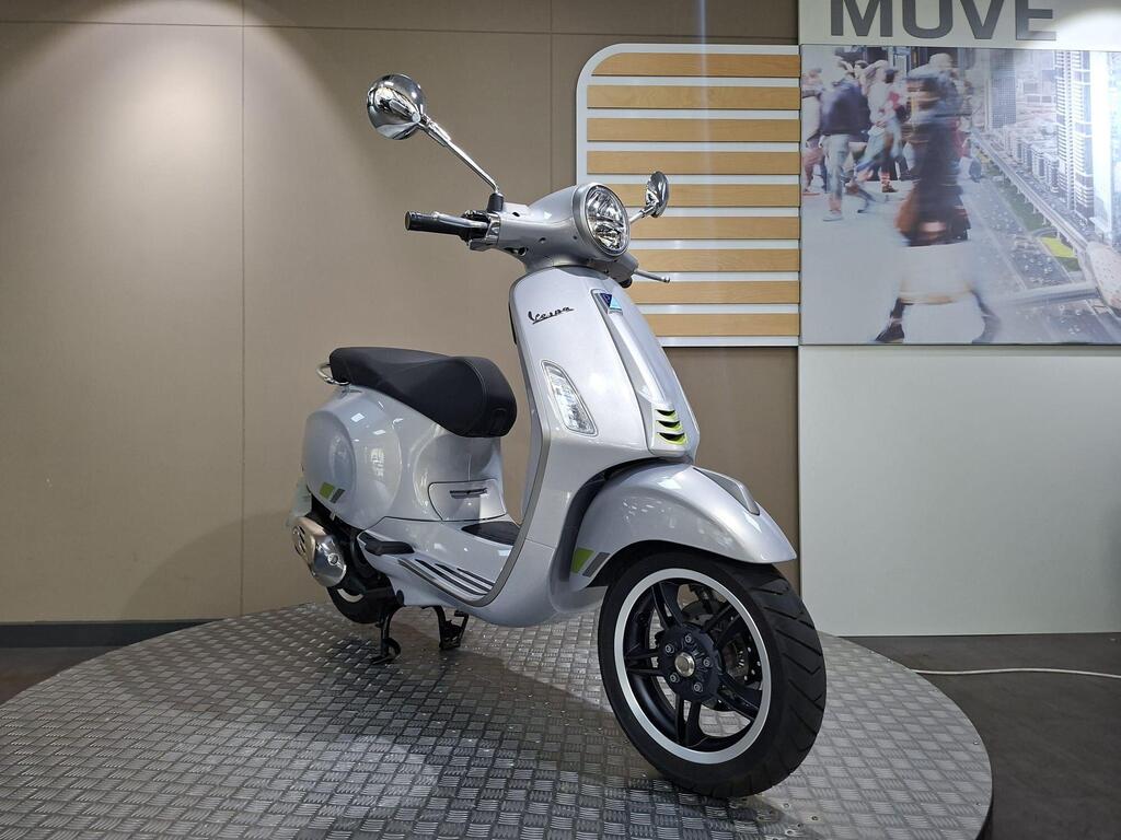 Vespa Primavera 125 2025 3 A sleek silver Vespa scooter is displayed on a circular platform, with a minimalist background featuring a cityscape image and the word "MOVE." The scooter showcases modern design elements, including chrome mirrors and a black seat.