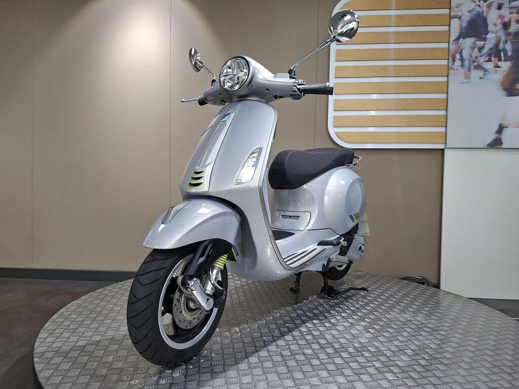 Vespa Primavera 125 2025 4 A silver scooter positioned at a slight angle on a textured platform, with a modern showroom backdrop featuring wooden paneling and a blurred crowd image.