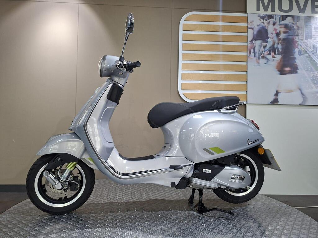 Vespa Primavera 125 2025 5 A sleek silver scooter displayed in a showroom, featuring a black seat and modern design elements, set against a neutral background with a blurred image of pedestrians.