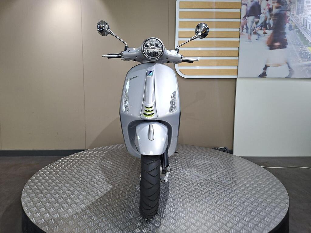 Vespa Primavera 125 2025 6 A silver scooter displayed on a circular platform with a textured surface, featuring a rounded front, chrome mirrors, and a modern design, against a neutral wall background.