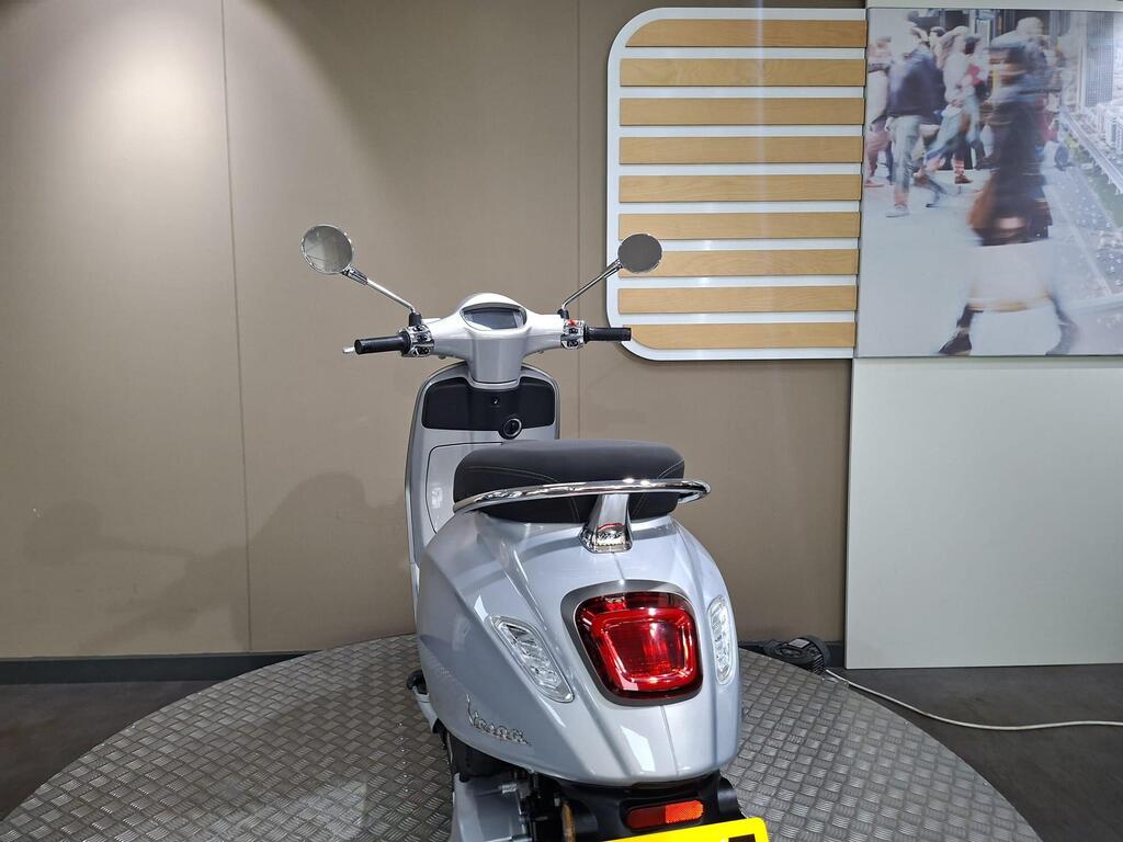 Vespa Primavera 125 2025 7 A silver scooter displayed in a showroom, viewed from the rear. The scooter features a sleek design with side mirrors and a rounded tail light. A light-colored wall with a wooden panel accent is visible in the background, alongside a blurred image of pedestrians.