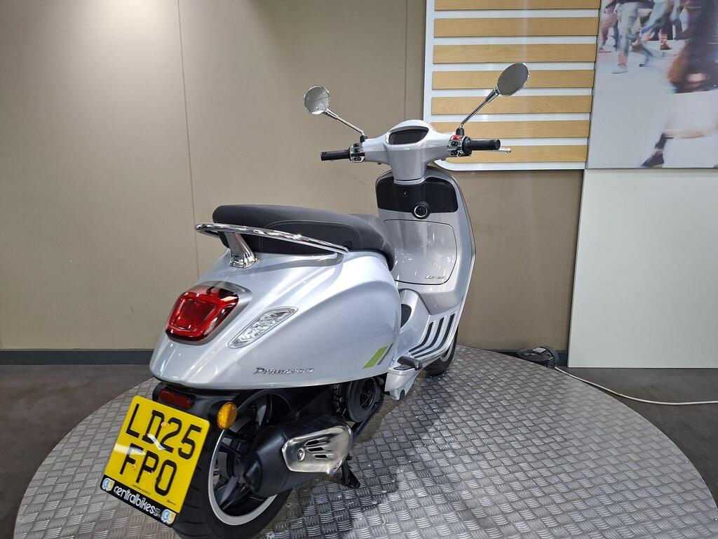 Vespa Primavera 125 2025 8 A silver scooter parked on a circular, textured platform, showcasing its rear view. The scooter's license plate is clearly visible, and a minimalist wall with simple stripes is in the background.