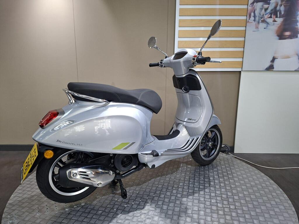 Vespa Primavera 125 2025 9 A sleek silver scooter is displayed on a circular metallic stand, showcasing its streamlined design with a black seat, rearview mirrors, and detailed branding on the side. The background features a minimalist design, emphasizing the scooter's modern aesthetics.