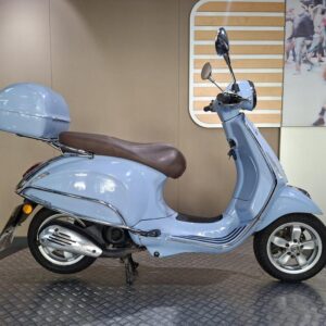 Vespa Primavera 50 2017 A blue scooter with a brown seat and a matching front storage compartment, displayed on a circular platform against a neutral background.