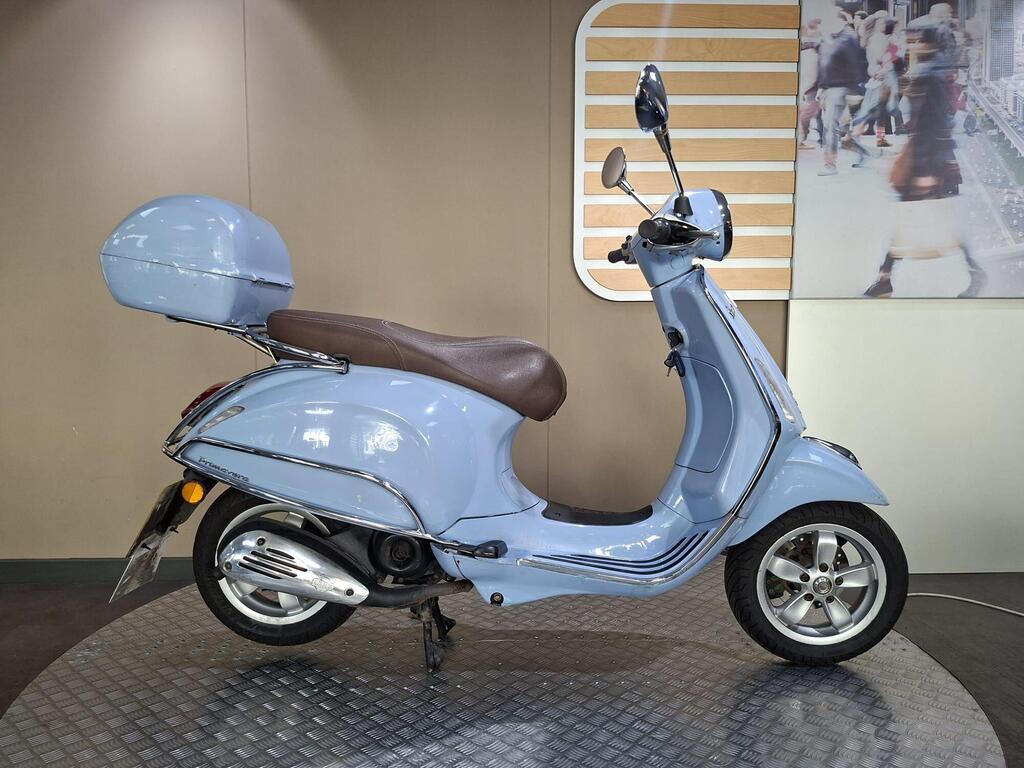 Vespa Primavera 50 2017 1 Vespa Primavera 50 2017 A blue scooter with a brown seat and a matching front storage compartment, displayed on a circular platform against a neutral background.