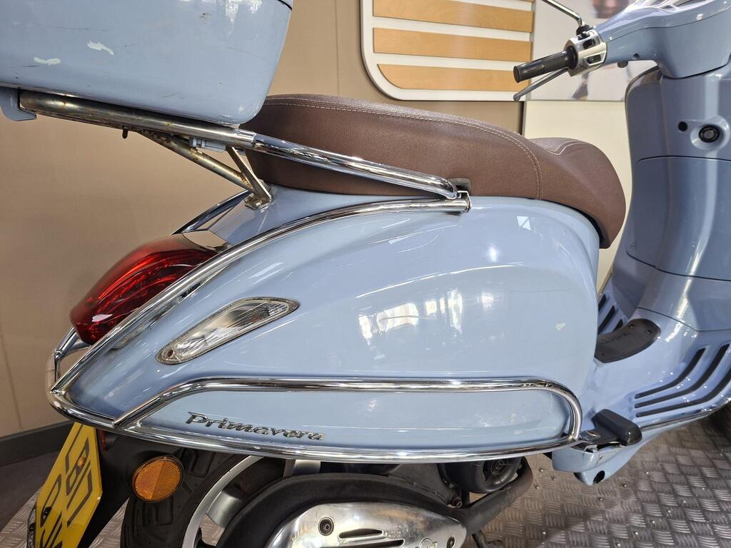 Vespa Primavera 50 2017 11 A close-up view of a light blue vintage scooter with a brown seat, showcasing its chrome detailing and a rear storage compartment. The scooter's name "Primavera" is visible on the side, and the background features a simple, modern interior.