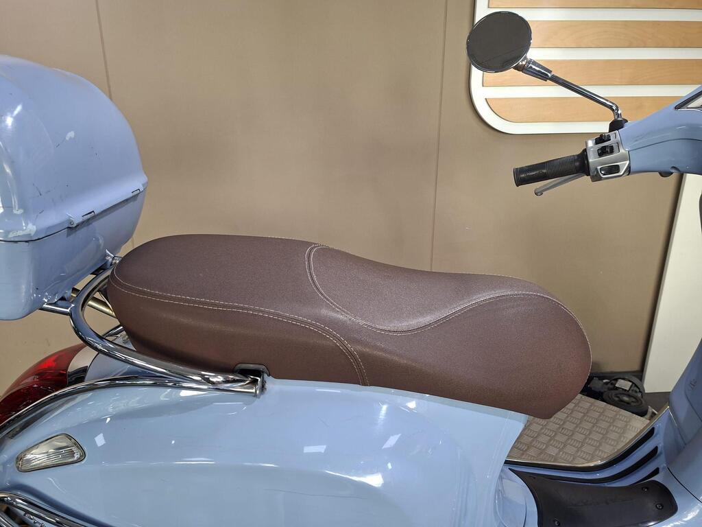 Vespa Primavera 50 2017 12 A side view of a light blue scooter with a brown seat, featuring a chrome grab rail and a rear storage box. The background is a neutral-colored wall.