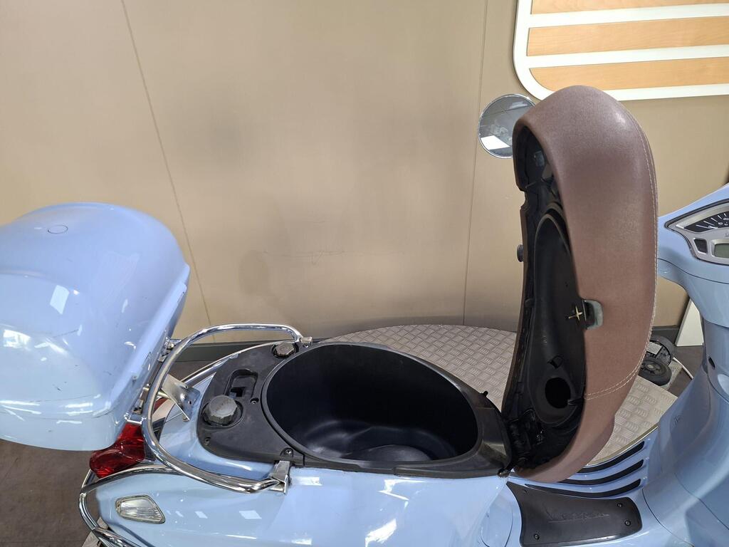 Vespa Primavera 50 2017 13 A close-up view of a blue scooter's rear storage compartment, with the lid opened, revealing the interior space. The scooter features a brown seat back and chrome accents. The background is a neutral wall with a light wooden panel.