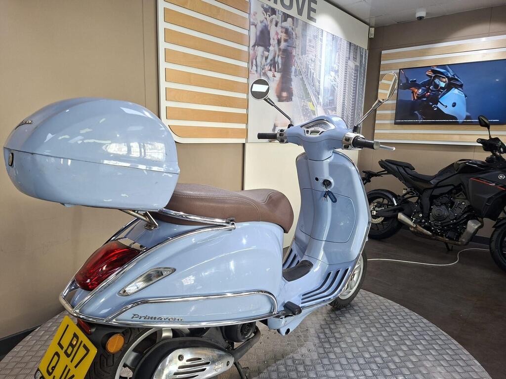 Vespa Primavera 50 2017 15 A light blue scooter with a brown seat and a rear storage compartment is displayed in a showroom, with a motorcycle visible in the background and branded signage on the walls.
