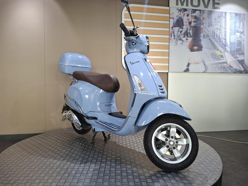 Vespa Primavera 50 2017 2 A light blue scooter with a brown seat, displayed on a textured circular platform in a showroom. The background features modern wall panels and an image of people in an urban setting.