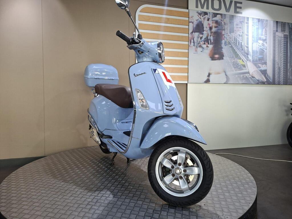 Vespa Primavera 50 2017 3 A pastel blue Vespa scooter displayed on a circular stand, featuring a brown seat and a learner's plate on the front, with a blurred urban background showcasing people and city buildings.