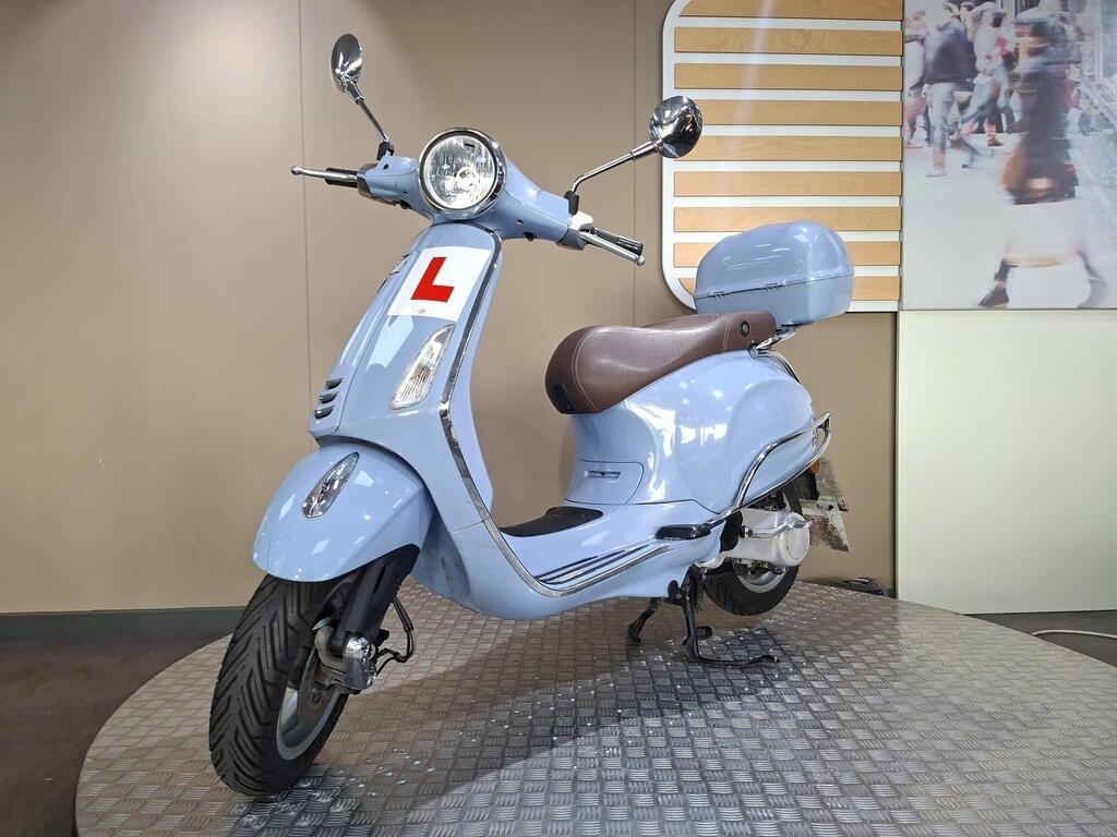 Vespa Primavera 50 2017 4 A light blue scooter with a brown seat displayed on a textured surface, featuring a learner's license plate marked with an "L," set against a neutral background with wooden accents.