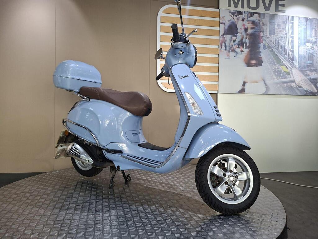 Vespa Primavera 50 2017 5 A light blue Vespa scooter with a brown seat and matching storage box, displayed on a textured platform against a neutral background.