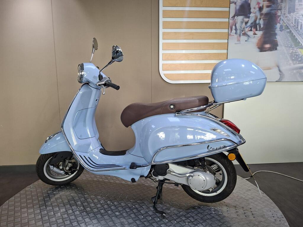Vespa Primavera 50 2017 6 A vintage-style light blue scooter with a brown seat and a rear storage box is positioned on a textured surface, set against a neutral-colored wall with a backdrop of a city scene.