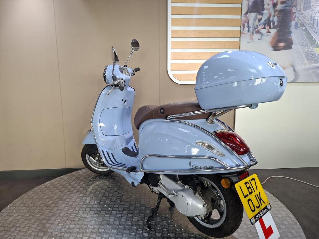 Vespa Primavera 50 2017 7 A light blue Vespa scooter with a brown seat and top box is displayed on a checkered surface, facing slightly to the left. The background features a neutral wall with a stylized image of a busy street scene.