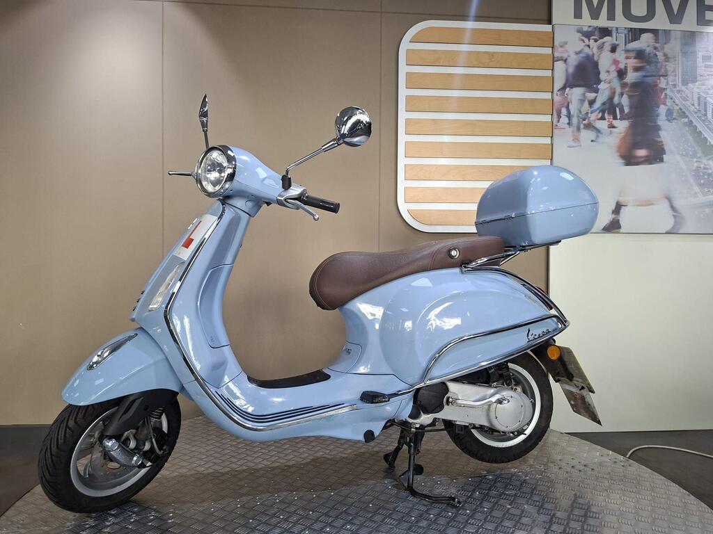 Vespa Primavera 50 2017 8 A light blue Vespa scooter parked on a textured surface, featuring a brown seat and a rear storage compartment, with a softly lit background showcasing a wall with modern design elements.