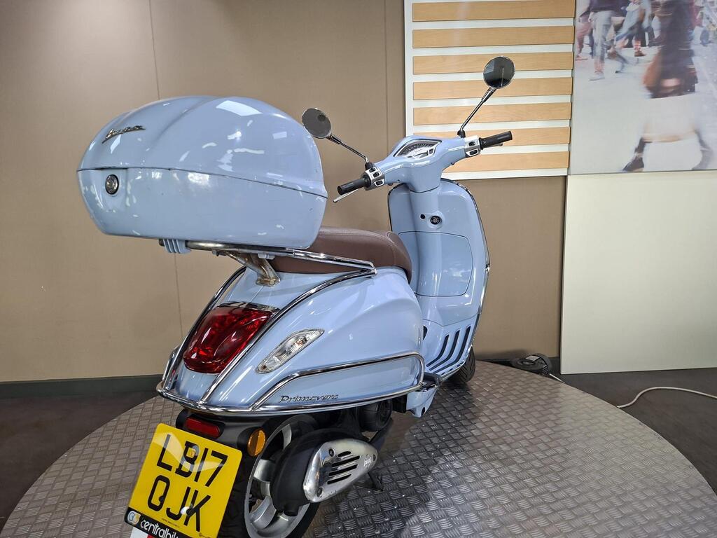 Vespa Primavera 50 2017 9 A light blue scooter with a brown seat and a rear storage compartment, displayed on a circular platform with a beige wall in the background.